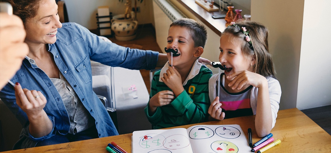 A woman smiles at two laughing children who are holding paper mustaches.