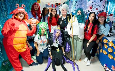 A group of MSK Kids staff members stand together, smiling, dressed as different characters from the Little Mermaid, in a hallway at MSK. 