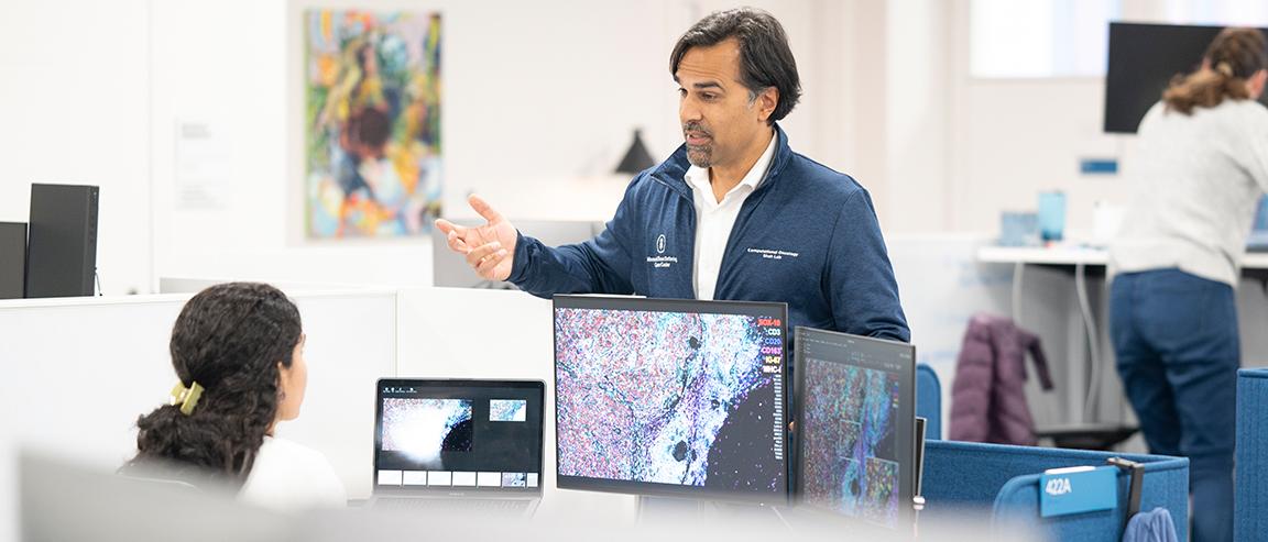 Dr. Sohrab Shah, Chief of Computational Oncology in the Department of Epidemiology and Biostatistics, speaks to an MSK staff member who is sitting in front of a computer monitor displaying an image of cells.