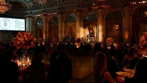 A dimly lit ballroom with elegant table settings and a screen projecting “The Society of Memorial Sloan Kettering.”