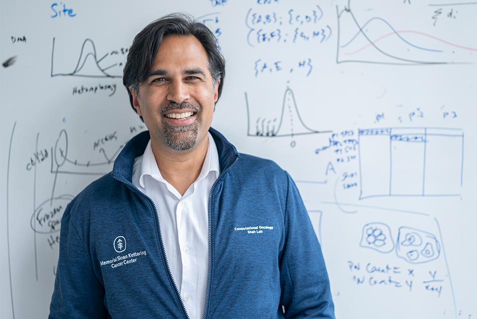 Dr. Sohrab Shah, standing in front of a white board with graphs on it and smiling. 