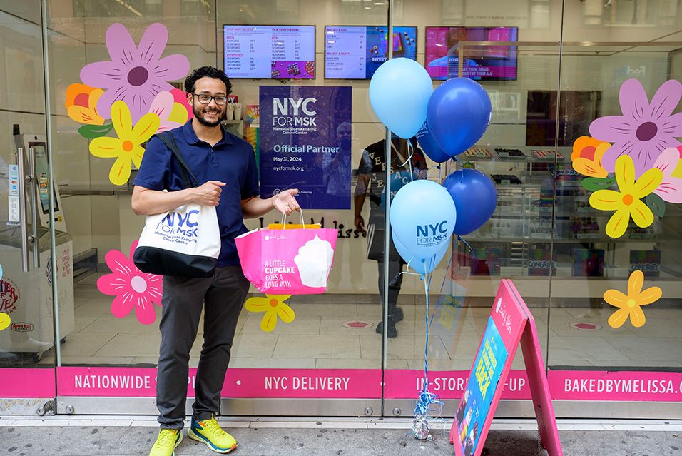 A young man holds a shopping bag with a cupcake logo on it. He stands in front of a glass door that displays an “NYC for MSK Official Partner” sign.  