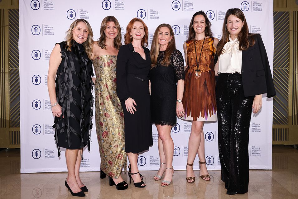 A group of six women in elegant attire stand in front of a step and repeat with The Society of MSK logo on it. 