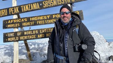 MSK donor John at Uhuru Peak in Tanzania. He is wearing a black parka and is standing in front of a sign that says "Africa's highest point."