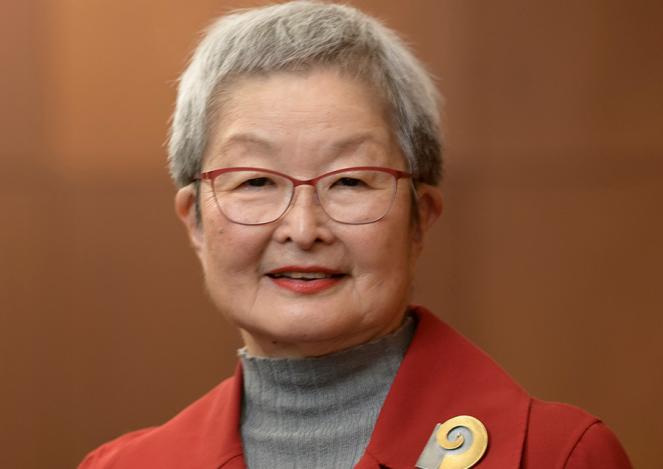 A woman with short gray hair in a gray mock turtleneck sweater and red blazer with a gold brooch stands, smiling, against a wood backdrop. 